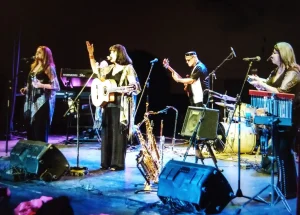 The band Sister Sister y Los Misters is setup on a stage. There are two speakers at the fron of the stage, one female guitarist, and one male guitarist in the image.